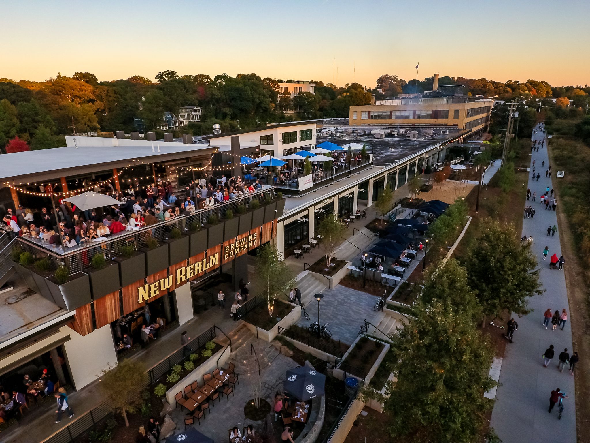 New Realm Brewing's rooftop is full of people and a sidewalk, the Beltline, shows pedestrians walking in both directions. The sun is fading from blue to orange.